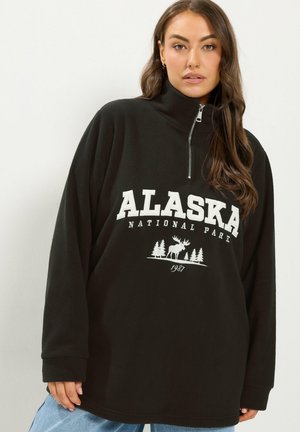 Femme portant un pull noir à fermeture éclair quart de longueur avec "Alaska National Park 1987" et un graphisme d'élan et d'arbres sur le devant.