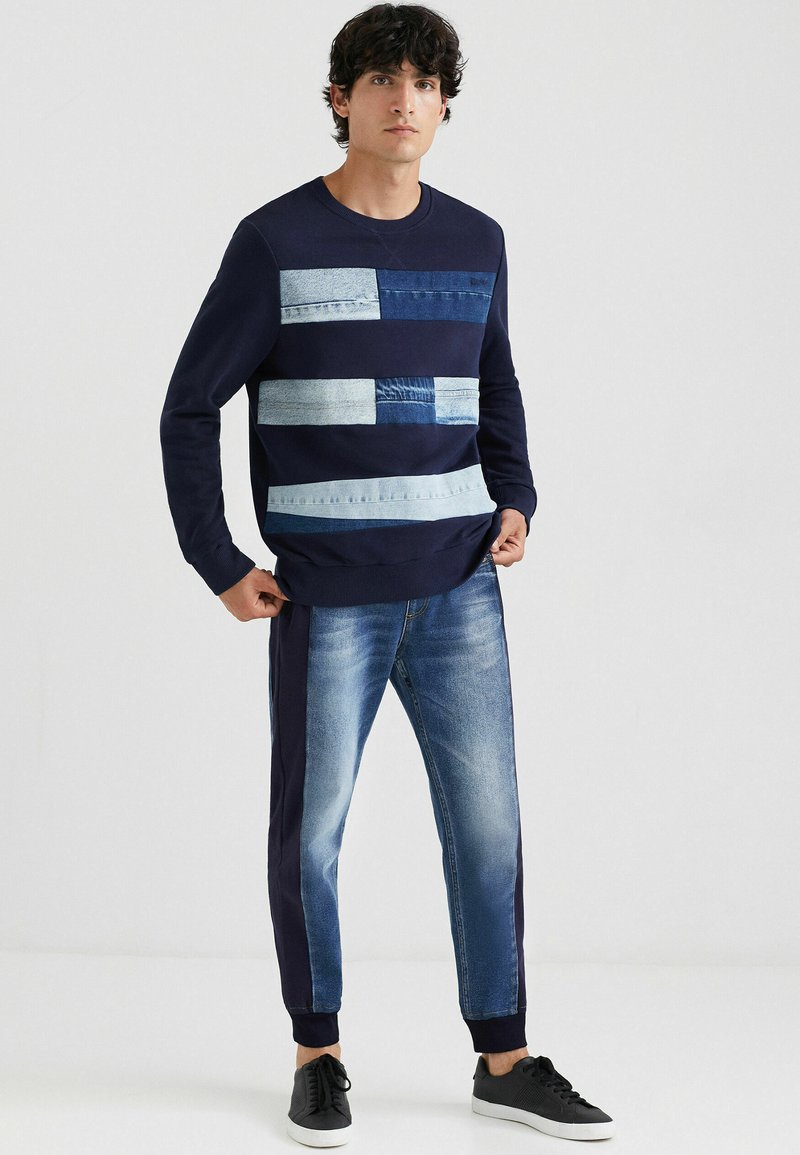 Homme portant un sweatshirt bleu marine avec un patchwork de denim clair et sombre, et un jean style jogger avec des panneaux latéraux noirs et des baskets noires.