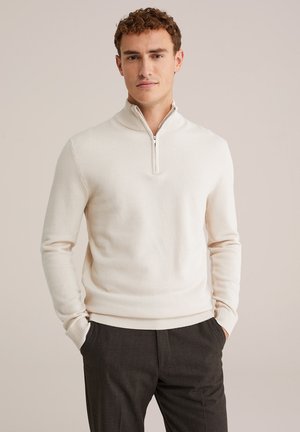 Sudadera de media cremallera color crema claro, confeccionada en un tejido de punto texturizado, con dobladillo y puños acanalados, combinada con pantalones oscuros.