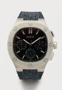 Ανδρικό ρολόι Guess με μαύρο καντράν με υφή, ασημένια δείκτες, υποκαντράν για ημερομηνία και ημέρα, ασημένιο στεφάνι με εμφανείς βίδες και λουράκι από υφασμάτινο μοτίβο.