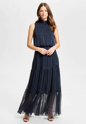 Femme aux longs cheveux portant une robe maxi bleu marine sans manches et des talons ouverts noirs, debout les mains jointes devant elle.