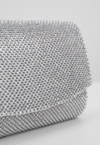 Pochette recouverte de petits strass argentés brillants avec un rabat de fermeture, présentée sur un fond clair uni.