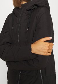 Chaqueta de plumas negra con capucha, que presenta un cierre frontal con cremallera y bolsillos laterales, textura suave y puños elásticos para mayor calidez.