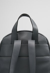 Sac à dos en cuir gris avec un sommet arrondi, deux bretelles et une texture lisse avec des coutures contrastées. Pas de détails matériels visibles.