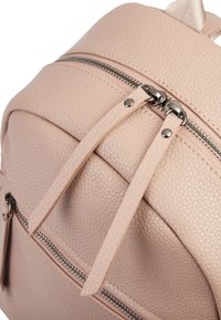 Zbliżenie na teksturowany, jasnoróżowy skórzany plecak z podwójnymi srebrnymi suwakami i długimi skórzanymi zawieszkami na suwakach.