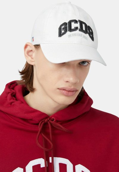 Młoda osoba nosząca białą czapkę GCDS i czerwoną bluzę GCDS z troczkami, patrząca w dół na jednolitym tle.