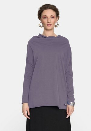 Kobieta o krótkich kręconych włosach, nosząca duże kolczyki w kształcie serca, luźną fioletową bluzkę z długim rękawem, czarną spódnicę oraz srebrną bransoletkę.