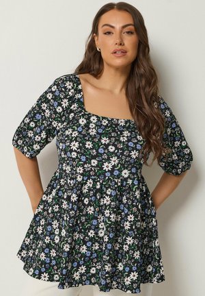 Femme aux longs cheveux bruns ondulés portant une robe noire à fleurs blanches et bleues, avec un décolleté carré et des manches bouffantes mi-longues jusqu'au coude.