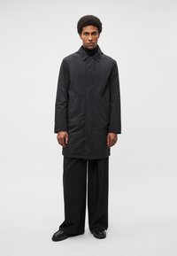 Fekete, vízálló kabát gallérral, egyenes szabású és oldalzsebes, fekete, bő szárú nadrág és csizma fölött viselve. Simogató textúra, minimalista kialakítás.