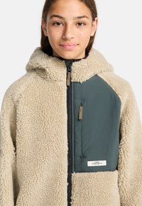 Giovane che indossa una giacca in pile sherpa beige con una tasca sul petto a zip verde scuro e cappuccio, rivolto in avanti.
