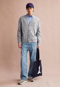 Cardigan gris, chemise à rayures bleues, jeans bleu clair, baskets grises, sac fourre-tout bleu marine avec logo blanc, porté par un modèle masculin sur un fond beige.