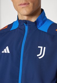 Giacca zip-up navy con accenti blu, caratterizzata da un'emblema bianca Adidas e dal logo del team, realizzata in tessuto morbido con colletto alto.