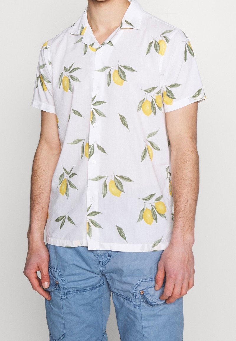 Homme portant une chemise à manches courtes blanche à motifs de citrons jaunes et de feuilles vertes, assortie à un short cargo bleu clair.
