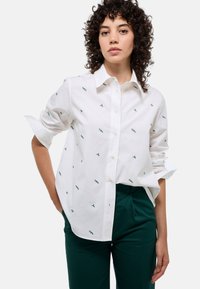 Chemise blanche à boutons avec des détails brodés verts, manches longues retroussées, associée à un pantalon vert. Comprend un col classique et une fermeture à boutons.