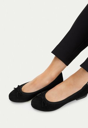 Pies con bailarinas negras con pequeños lazos, combinadas con pantalones negros recortados sobre un fondo blanco.