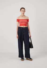 Crop top off-the-shoulder a righe rosse e bianche, abbinato a jeans a gamba larga blu scuro. La modella tiene una piccola borsa nera. Sneakers con suole in gomma.