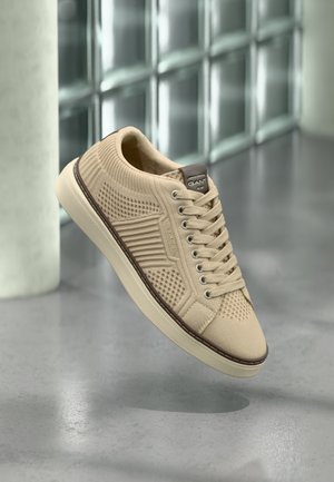 Bézs sportcipő struktúrált kötött felsőrésszel, kerek orral és lapos fűzőkkel. Kontrasztos barna sarokrész és gumitalp, tapadós mintával.