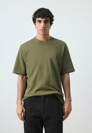 Junger Mann mit lockigem dunklem Haar, trägt ein olivgrünes kurzärmeliges Hemd und schwarze Jeans, steht vor einem schlichten hellen Hintergrund.