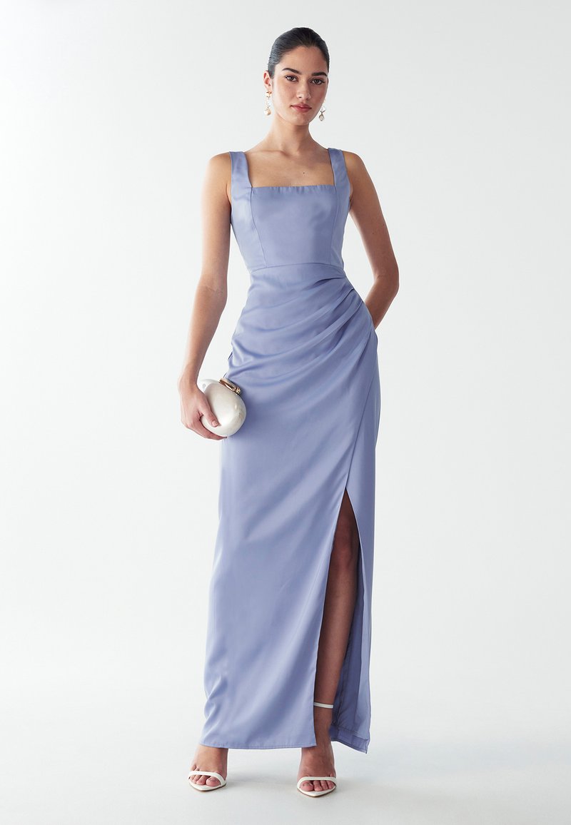 Długa lawendowa satynowa sukienka z kwadratowym dekoltem, bocznym rozcięciem i marszczonym teksturem. Uzupełniona białymi szpilkami i małą okrągłą kopertówką.
