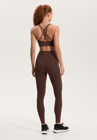 Rjava športna modrček z zarezovanimi naramnicami na hrbtu, združen z rjavimi visokim pasom legicami. Modelka nosi črne športne čevlje. Gladek teksturen material.