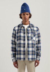 Knoop-up jack van fleece, met een blauw, wit en grijs geruit patroon. Inclusief een borstzak en een wit T-shirt met ronde hals eronder.