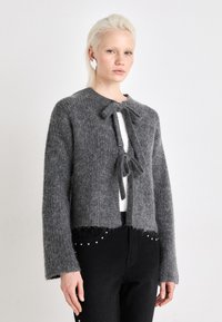 Szürke kötött kardigán, cropped fazonban, nyitott elől és masni zárással. Puha textúra, rojtos szegéllyel. Fehér felső fölött viselve.