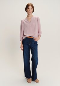 Donna che indossa una blusa rosa chiaro, jeans a gamba larga blu scuro con una cintura marrone e sandali beige aperti davanti, in piedi con una mano in tasca.