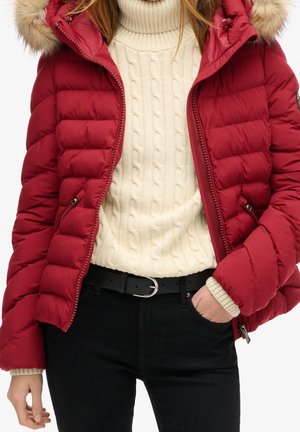 Czerwona puchowa kurtka z kapturem obszytym futerkiem, kremowy sweter w warkocze, czarny pasek i czarne spodnie, ręka w kieszeni.