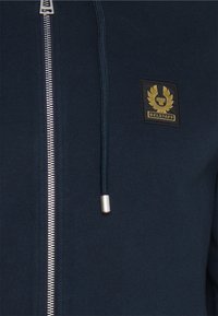 Marinblå zip-up hoodie med silverfärgad dragkedja, snörning med metallspets och guldig Belstaff-logotyp på vänster bröst.