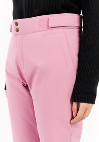 Roze broek heeft een gladde, matte textuur, twee bronzen knopen bij de taille en een patchzak aan de zijkant. Het merklogo is zichtbaar.