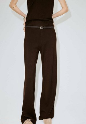 Persona con pantalones anchos de pierna en marrón oscuro, con un cinturón delgado oscuro y zapatos beige, de pie con las manos en las caderas contra un fondo liso.