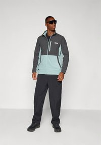 Hall ja hele sinine lukuga jakk rinnataskuga, kombineeritud mustade pükste ja mustade kingadega. Modell kannab tumedaid päikeseprille.