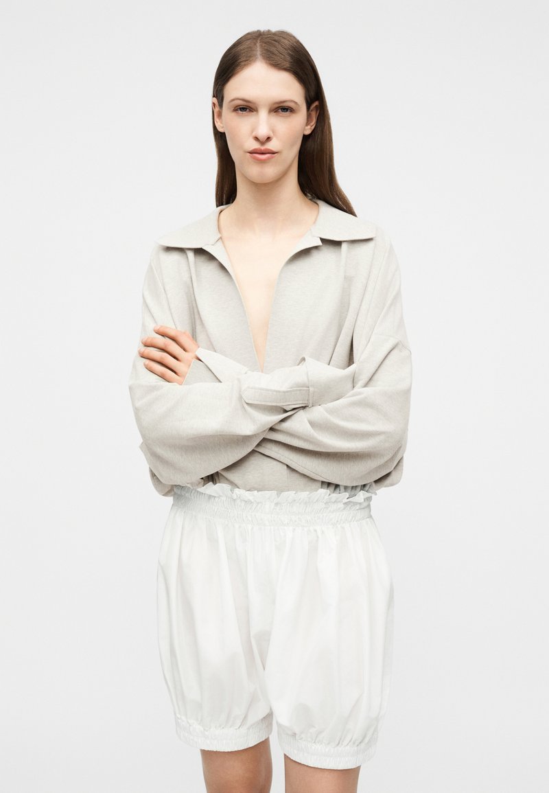 Femme aux longs cheveux bruns portant un haut à manches longues beige clair et un short blanc ample, les bras croisés, fond neutre.