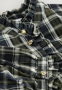 Bluzka s kostkovaným vzorem v zelené a námořnické modři, zdobená volánovým límcem a zlatými knoflíky, vyrobena z měkké látky s texturovaným povrchem.