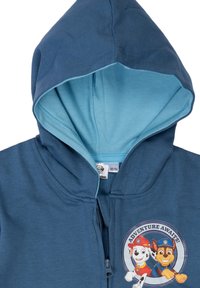 Felpa blu con zip e fodera azzurro chiaro, caratterizzata da una grafica circolare "Adventure Awaits" con personaggi di cani in stile cartoon sul davanti.