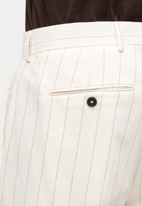 Κοντινό πλάνο λευκών παντελονιών με λεπτές κάθετες καφέ ρίγες, με μαύρη πίσω τσέπη με κουμπιά και θηλιές για ζώνη.