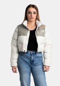 Giacca cropped imbottita in bianco e grigio chiaro, con colletto alto, chiusura a zip e logo sul lato sinistro del petto. Indossata con jeans a vita alta.