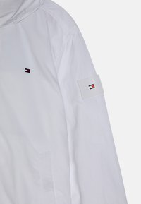 Biała, lekka kurtka z kapturem, z małym wyszywanym logo i łatą z logo na rękawie. Gładka tekstura, prosty design.