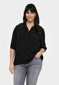 Camicia nera a bottoni con una trama morbida, caratterizzata da un colletto plissettato, maniche corte e dettagli decorativi sulle spalle. Indossata con jeans grigi.