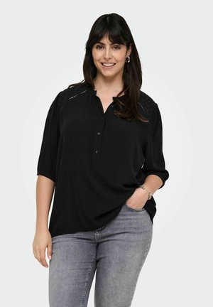 Chemise noire à manches courtes en tissu doux, avec un col plissé et des détails décoratifs aux épaules. Portée avec un jean gris.