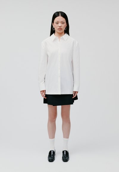 White button-up shirt with long sleeves, paired with a black mini skirt, white socks, and black loafers. Clean lines and minimal design.