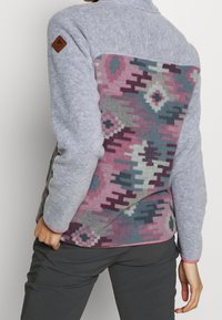 Pullover in pile con parte anteriore grigia e retro decorato in tonalità di rosa, blu e grigio; include un'etichetta marrone sulla spalla.