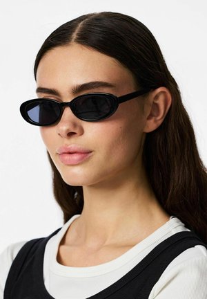 Nuori nainen, jolla on pitkät tummat hiukset ja joka käyttää kapeita mustia aurinkolaseja ja mustaa hihatonta yläosaa valkoisen paidan päällä, neutraali tausta.