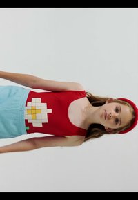 Niña con una camiseta sin mangas roja con un diseño pixelado de una flor blanca y amarilla y una falda azul clara, posando frente a un fondo gris liso.