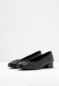 Black ballet flats made of smooth leather, featuring a shiny patent toe cap, small bow detail, and a low block heel.