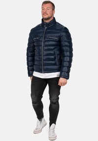 Giacca imbottita blu navy con colletto alto, tasche con zip e dettagli a costine. Indossata sopra una camicia bianca e abbinata a jeans neri strappati e sneaker bianche.