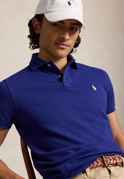 Junger Mann mit dunklem lockigem Haar, der eine weiße Kappe und ein blaues Polohemd trägt, sitzt auf einem Stuhl vor einem schlichten Hintergrund.