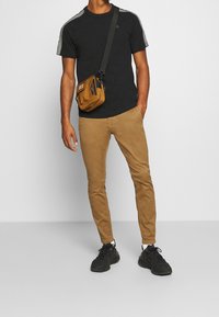 Black short-sleeve t-shirt with gray accents, paired with tan slim-fit chinos and black sneakers. A small brown crossbody bag is worn.