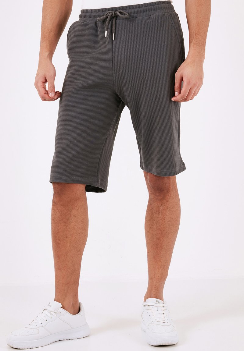 Grå træningsshorts med et struktureret, ribbet mønster. Har en elastisk talje med snoretræk, der slutter ved knæet. Hvide sneakers.