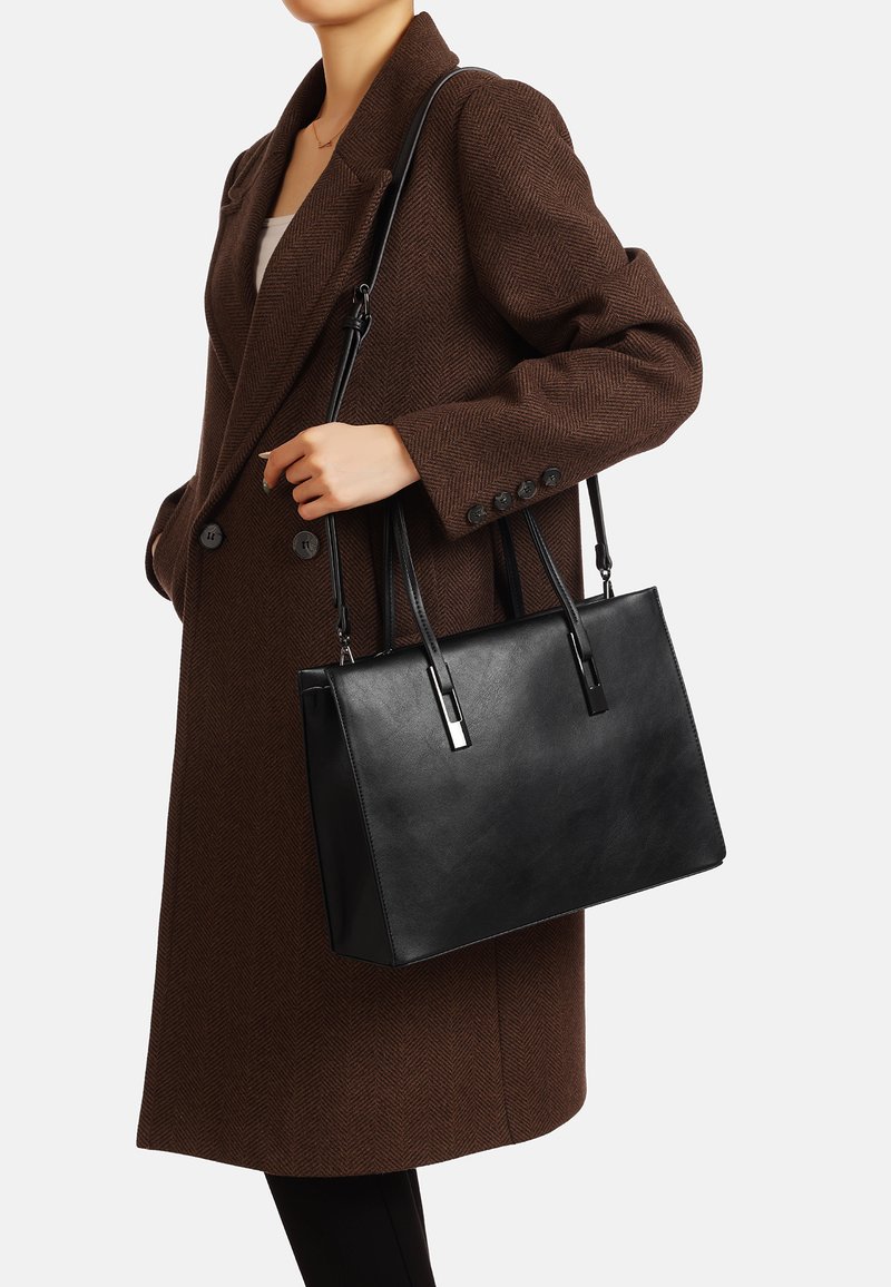 Czarna skórzana torba typu tote o strukturalnym kształcie, płaskim dnie, dwóch ramiączkach oraz metalicznymi srebrnymi akcentami. W zestawieniu z brązowym płaszczem w jodełkę.